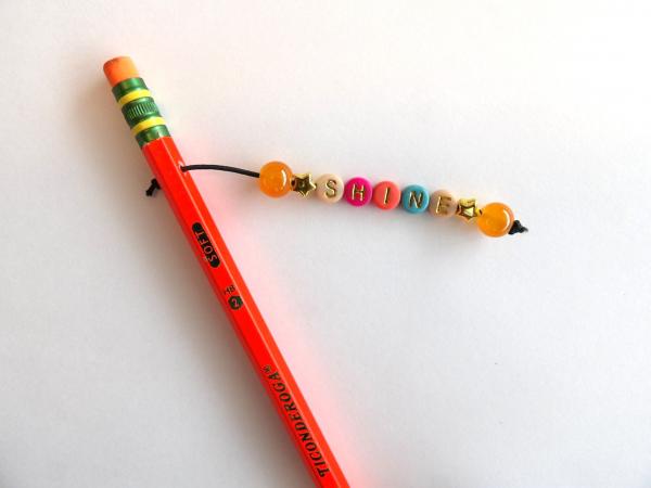 An orange pencil with a beaded charm with the word "Shine."