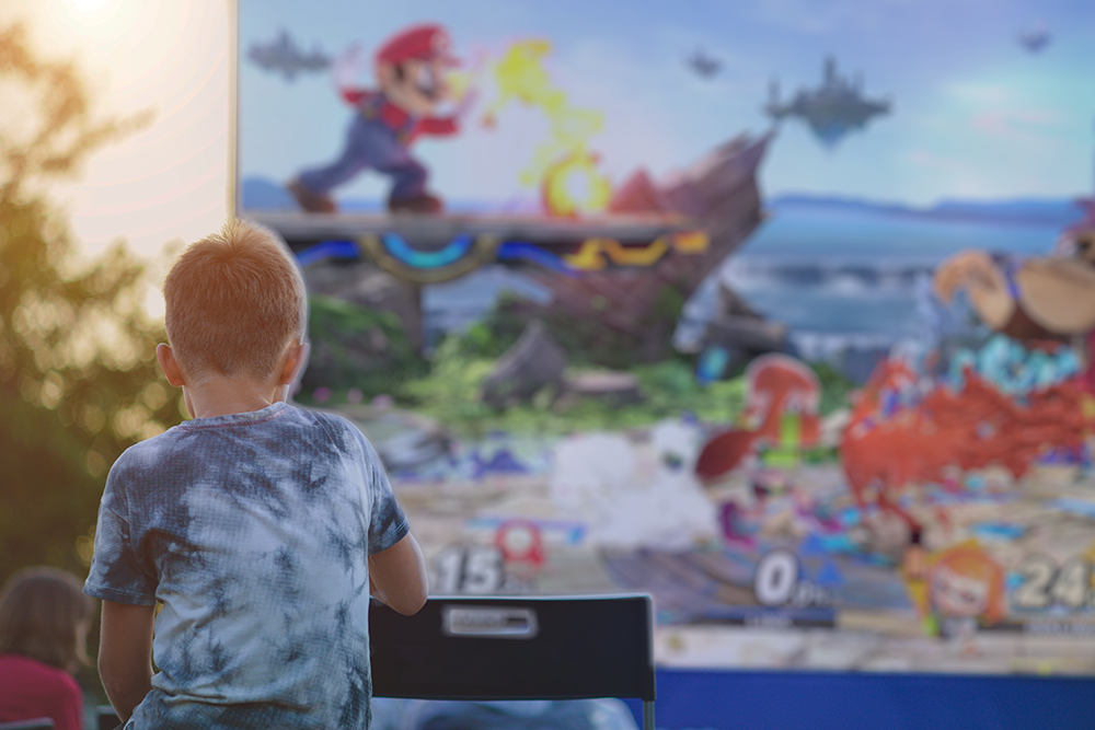 Young boy sitting in front of a Video game on a jumbo outdoor screen at dusk.