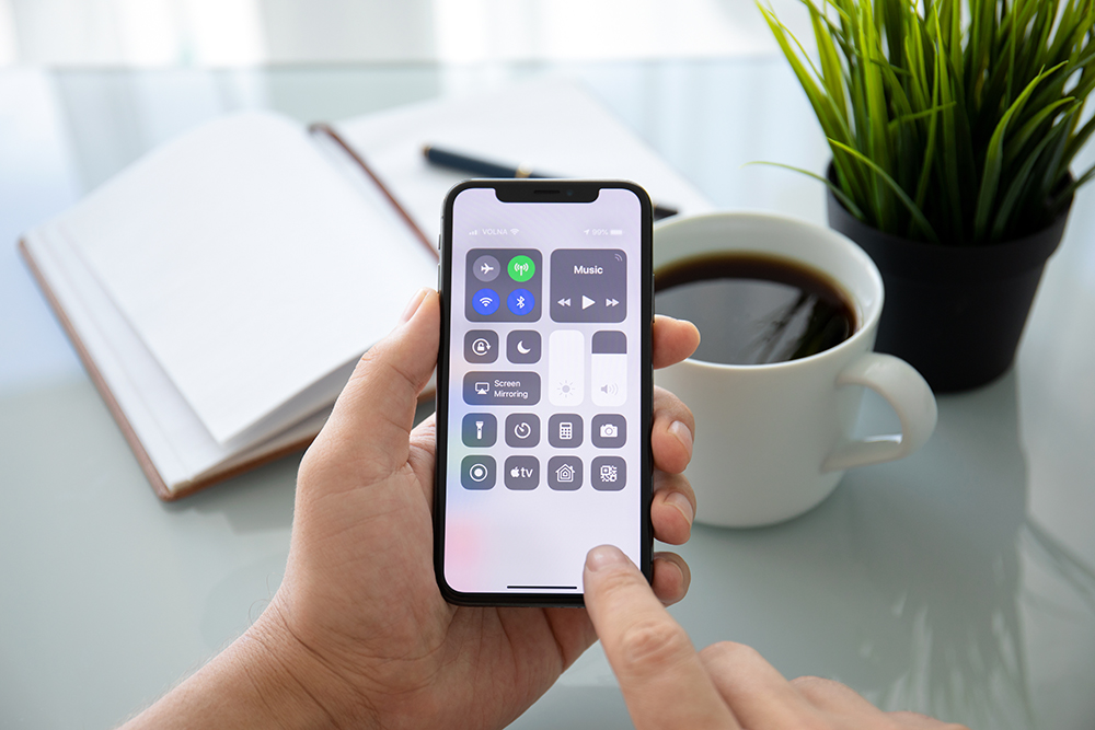 Hands holding an iphone showing the control center features. A notebook,  cup of coffee, and a fake plant are on a glass-top desk in the background.